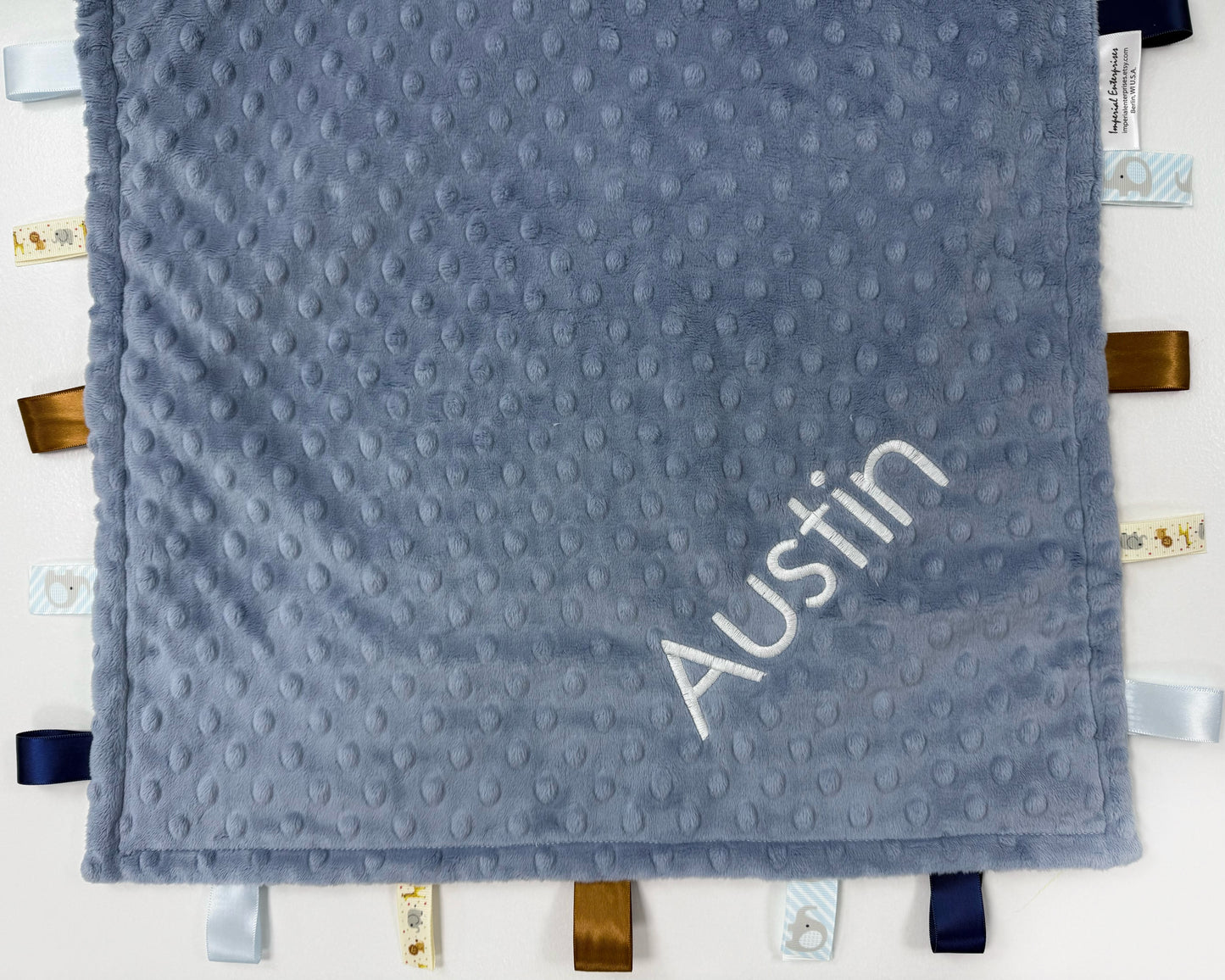 Gray textured blanket with 'Austin' embroidered on it, hanging on a white wall.