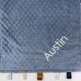 Gray textured blanket with 'Austin' embroidered on it, hanging on a white wall.