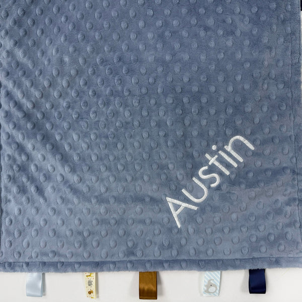 Gray textured blanket with 'Austin' embroidered on it, hanging on a white wall.