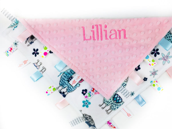 A personalized ribbon tag blanket with pink minky fabric, featuring the name 'Lillian' embroidered in pink, with a pattern of pink and teal llamas, and multiple ribbons on the edges.
