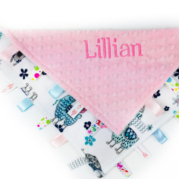A personalized ribbon tag blanket with pink minky fabric, featuring the name 'Lillian' embroidered in pink, with a pattern of pink and teal llamas, and multiple ribbons on the edges.