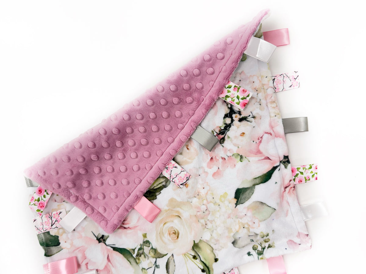 Floral-patterned tag blanket with pink textured border and ribbons on a white background